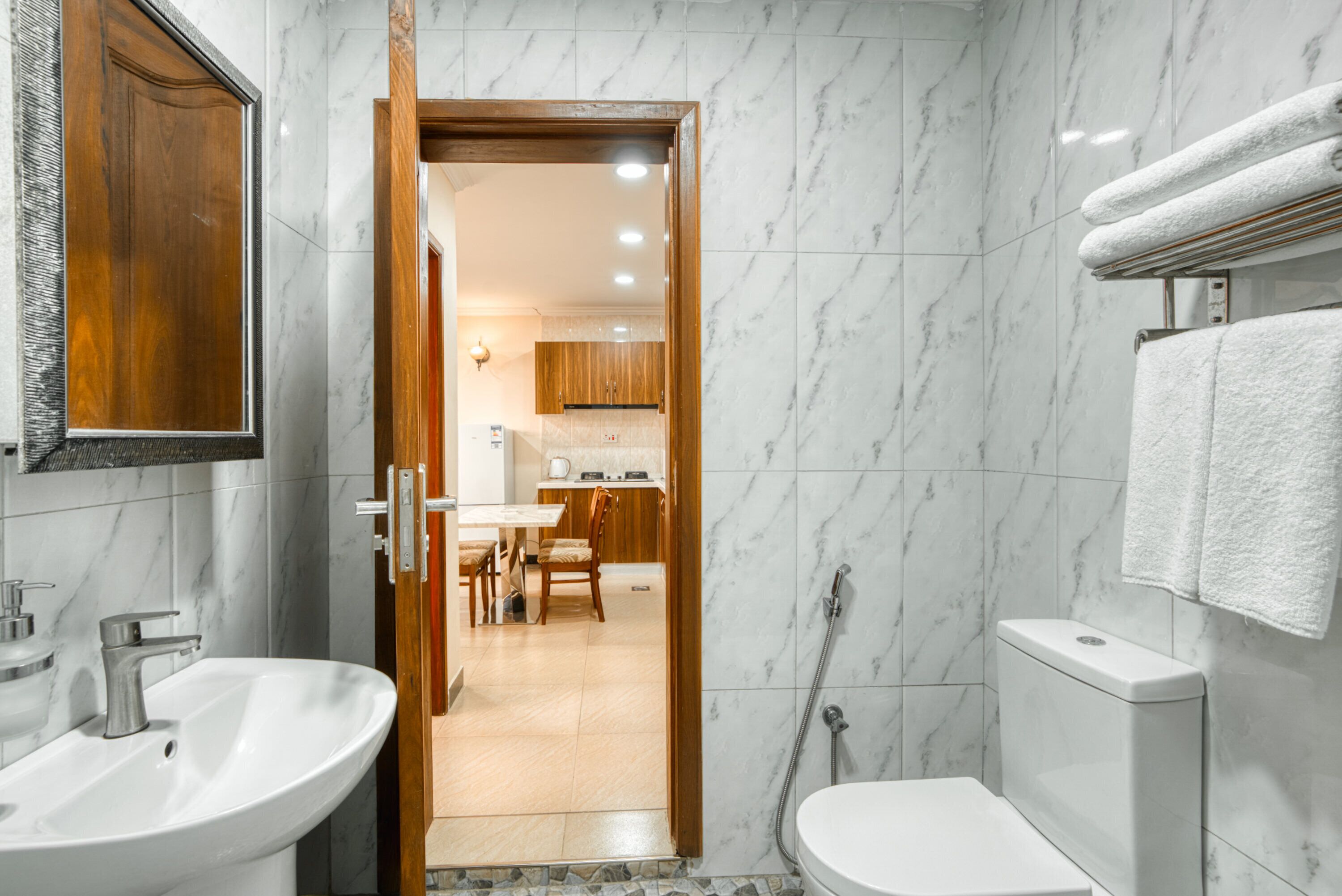 Baño con lavamanos, inodoro y ducha. Una puerta de madera da a un comedor con mesa y sillas.