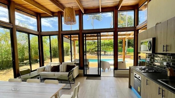 A spacious kitchen with large windows, a dining area with white furniture, and a view of a swimming pool.