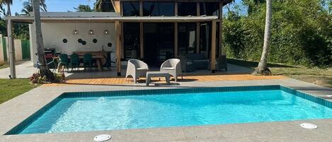 Una piscina con sillas de descanso, un área de terraza cubierta y palmeras al fondo.