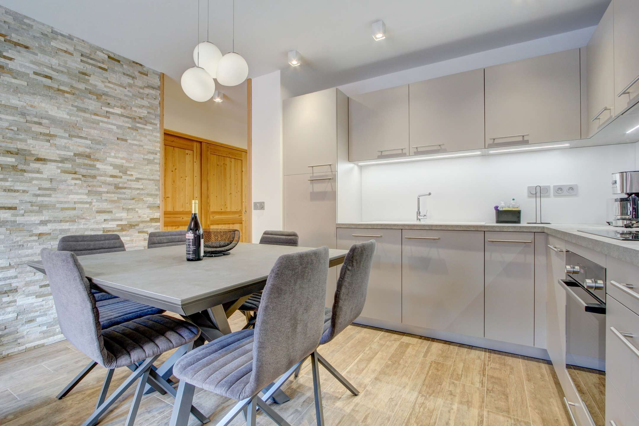 Een moderne keuken met grijze kastjes, een houten deur en een eethoek met een tafel en stoelen.