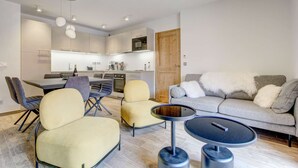 A modern living room with a sofa, two armchairs, and a coffee table. There is a kitchen in the background with white cabinets and a wooden door. - Carlina 203 Beautiful ski in ski out 2 bed + cabin  in the heart of the village (MORZINE)