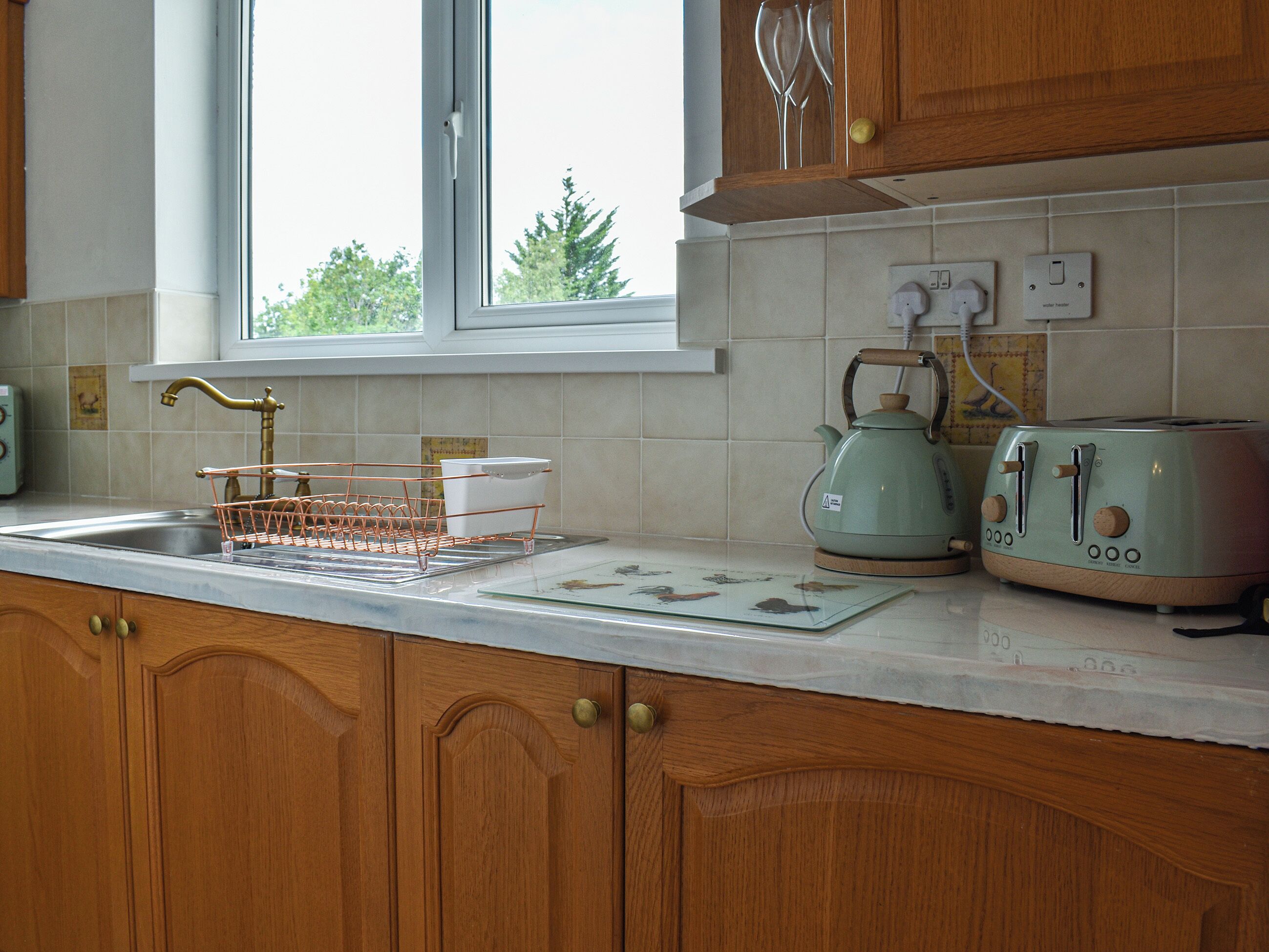 A kitchen with wooden cabinets, a window, a sink, a kettle, and a toaster.