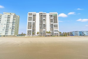 A modern high-rise building with multiple balconies and a flat roof, situated next to a sandy beach with palm trees.
