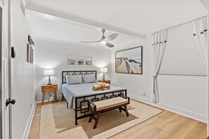 Quarto com cama, mesas de cabeceira, um banquinho, ventoinha no teto e uma pintura com tema praiano.