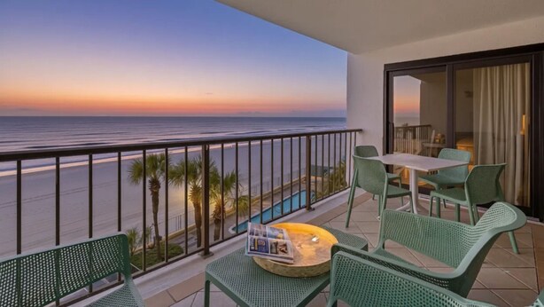 A balcony with a view of the ocean at sunset, green wicker furniture, and a small table with a magazine.
