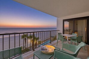 Uma varanda com vista para o oceano ao pôr do sol, mobiliário de vime verde e uma pequena mesa com uma revista.