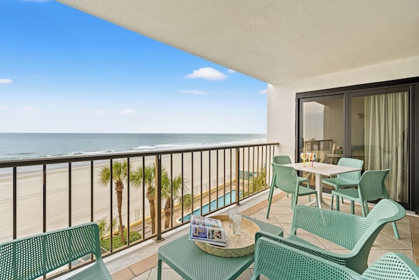 A balcony with a view of the beach and ocean, green wicker furniture, and a table with a magazine and glass.