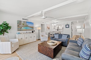 A spacious living room with a large flat-screen TV, a coffee table, a sofa, and a striped armchair. There is a dining area with a table and chairs, and a kitchen in the background.