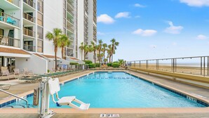 A swimming pool adjacent to a high-rise building with balconies, palm trees, and a beach view.