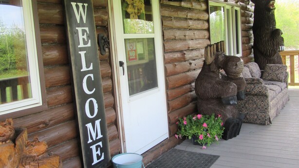 Une cabane en bois avec une pancarte « BIENVENUE », une statue d’ours et une véranda dotée d’un banc et de plantes en pot.