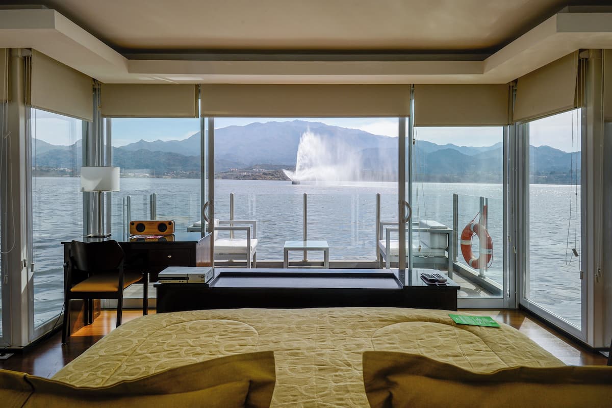 Una habitación de hotel moderna con una cama grande, un escritorio y vistas a un lago con montañas y una fuente.
