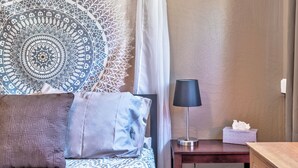 A neatly arranged bedroom with a patterned tapestry, a bedside lamp, and a bedside table with a tissue box. - Red Rocks, HOT TUB, W. Sedona, Walk to trls, 1 bd! (Sedona)