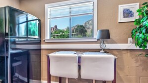 A kitchen with a refrigerator, a window with blinds, a table with two chairs, and a plant. - Red Rocks, HOT TUB, W. Sedona, Walk to trls, 1 bd! (Sedona)