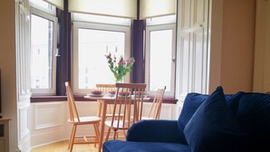 Un salon avec un canapé bleu, des chaises en bois, une table à manger avec de l’assiette, et une fenêtre donnant sur des immeubles.