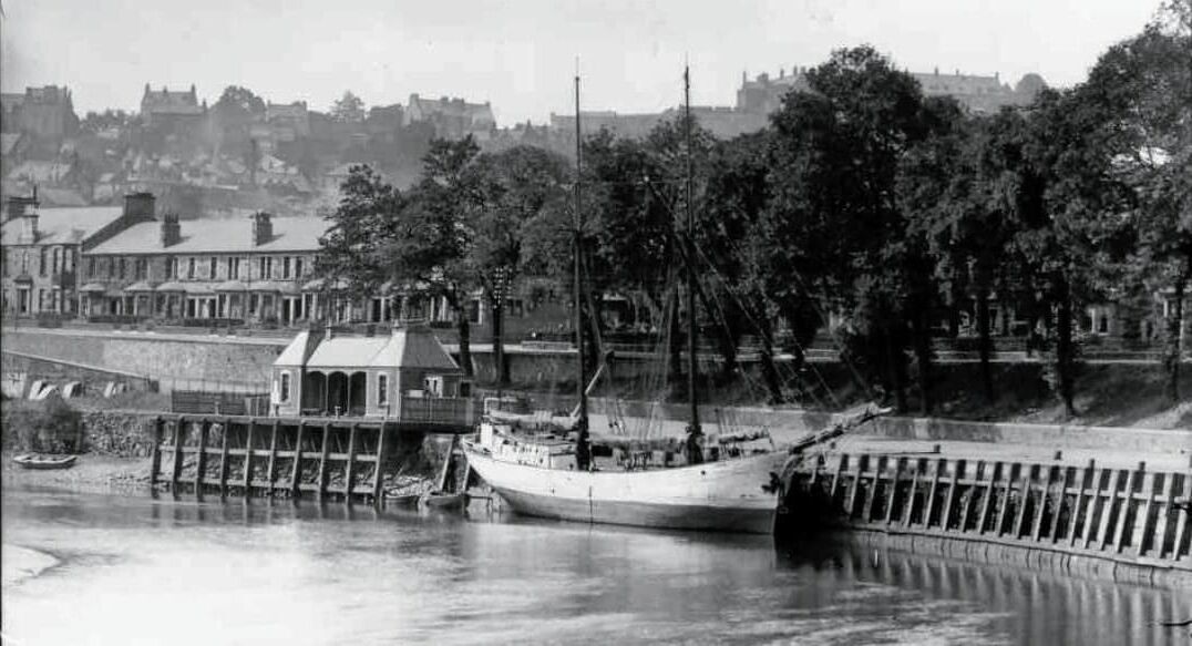 A historic waterfront scene with a large sailing ship docked at a pier, surrounded by trees and buildings.