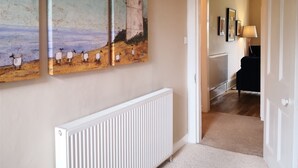Un couloir avec un radiateur blanc, un tableau représentant des moutons sur une plage et une porte donnant sur une autre pièce.