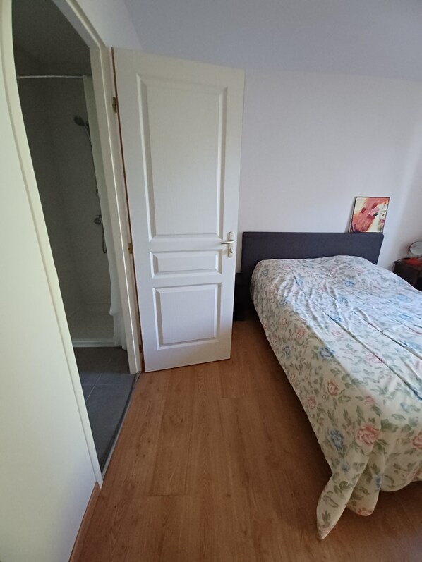 Ein Schlafzimmer mit Holzboden, einem Bett mit floralem Überwurf, einer weißen Tür und einem sichtbaren Duschbereich durch eine offene Tür.