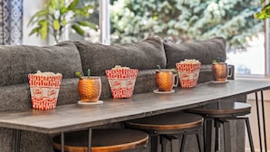 A row of popcorn containers and copper mugs on a table in a cozy seating area with a view of greenery outside.