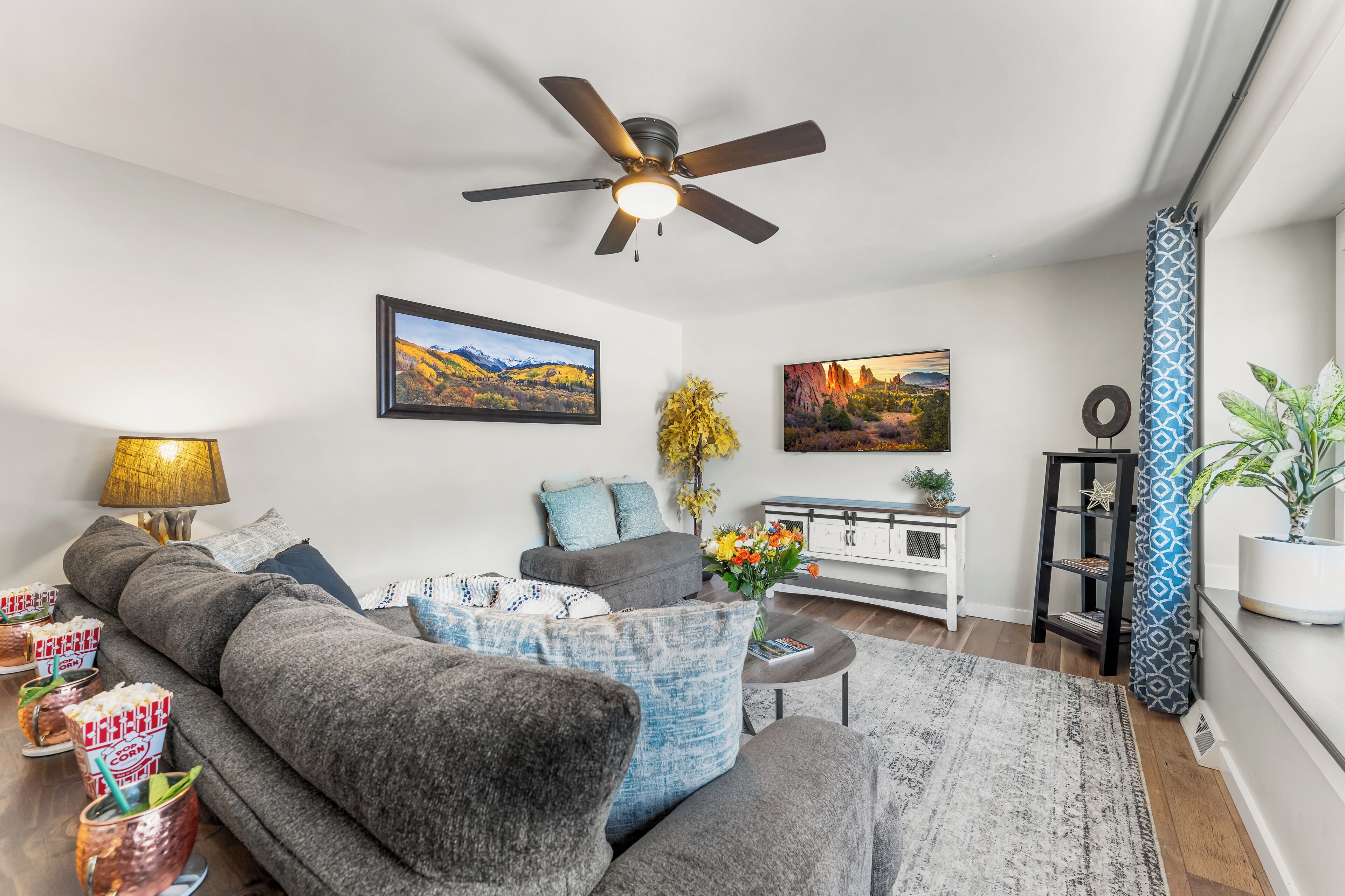 A modern living room with a grey sofa, a ceiling fan, and two framed landscape paintings.