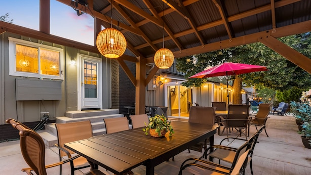 An outdoor patio with a wooden table and chairs, surrounded by a covered area with hanging wicker lamps.