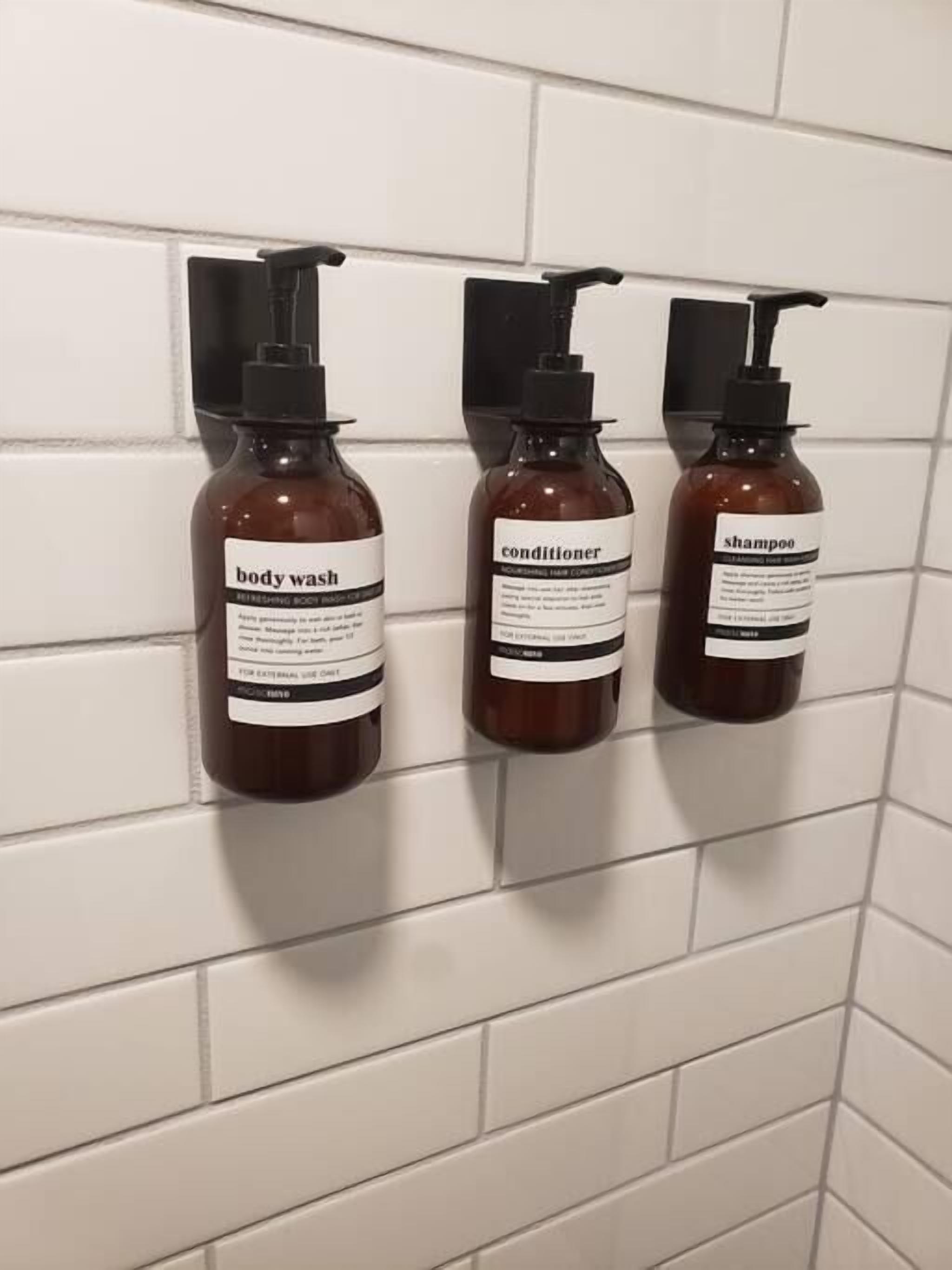 Trois bouteilles de gel douche, d’après-shampoing et de shampoing sont suspendues à un mur recouvert de carreaux blancs.