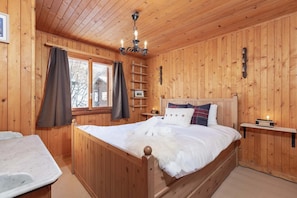 Quarto com painéis de madeira, uma cama, mesa de cabeceira, prateleira e janela com cortinas.