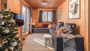 A cozy cabin interior with a wooden interior, a gray sofa with patterned cushions, a Christmas tree, and a framed animal head on the wall.