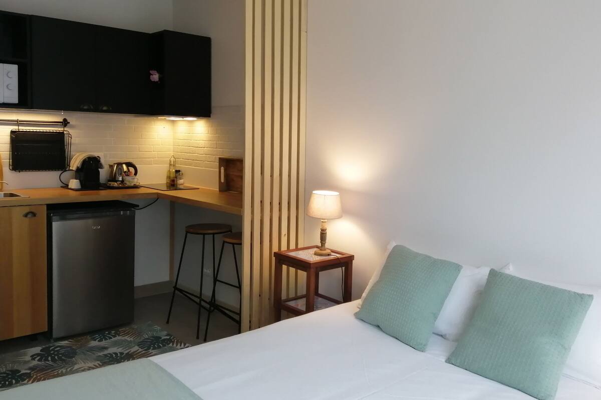 A compact hotel room with a kitchenette, a bed with green pillows, a small table with a lamp, and a patterned rug.