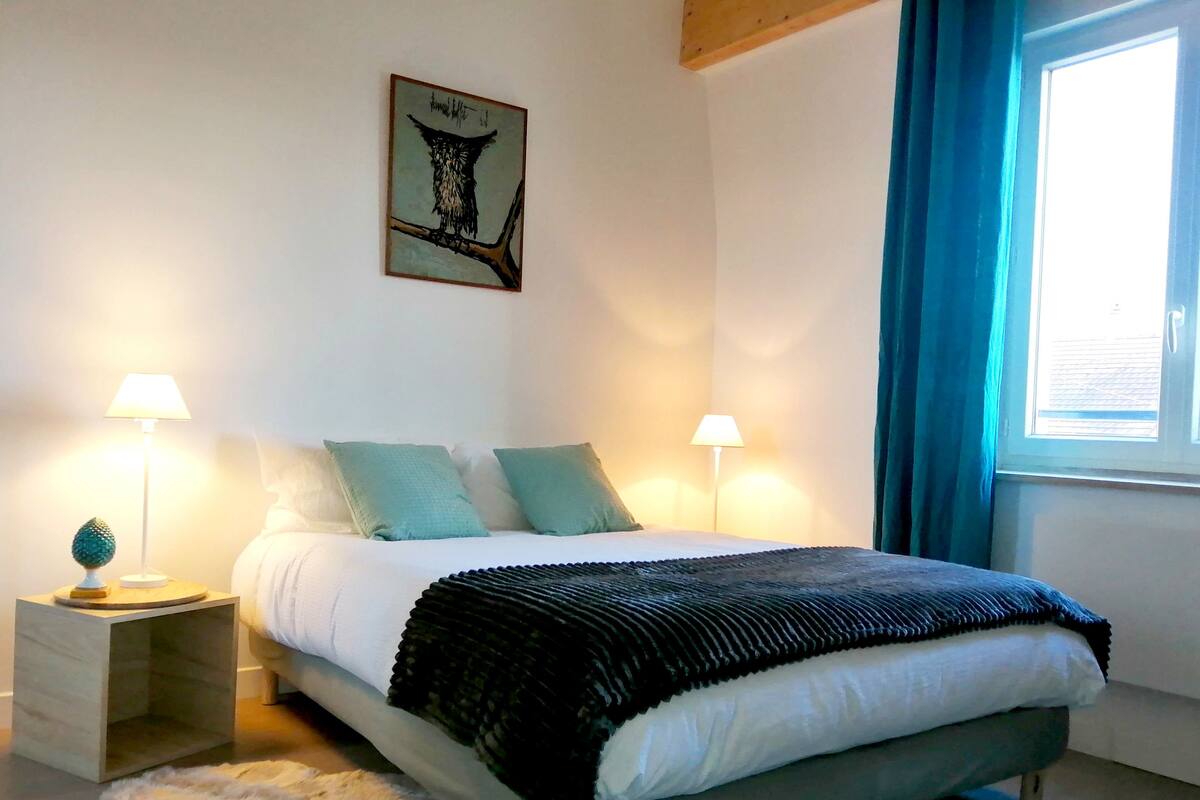 A bedroom with a bed, two bedside tables, a lamp, a fluffy rug, a window with curtains, and a wall art piece.