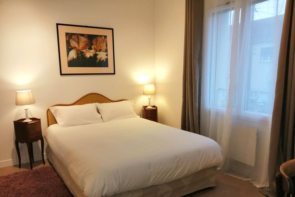 A neatly made bed with white linens, two bedside tables with lamps, a framed picture on the wall, and a window with curtains.