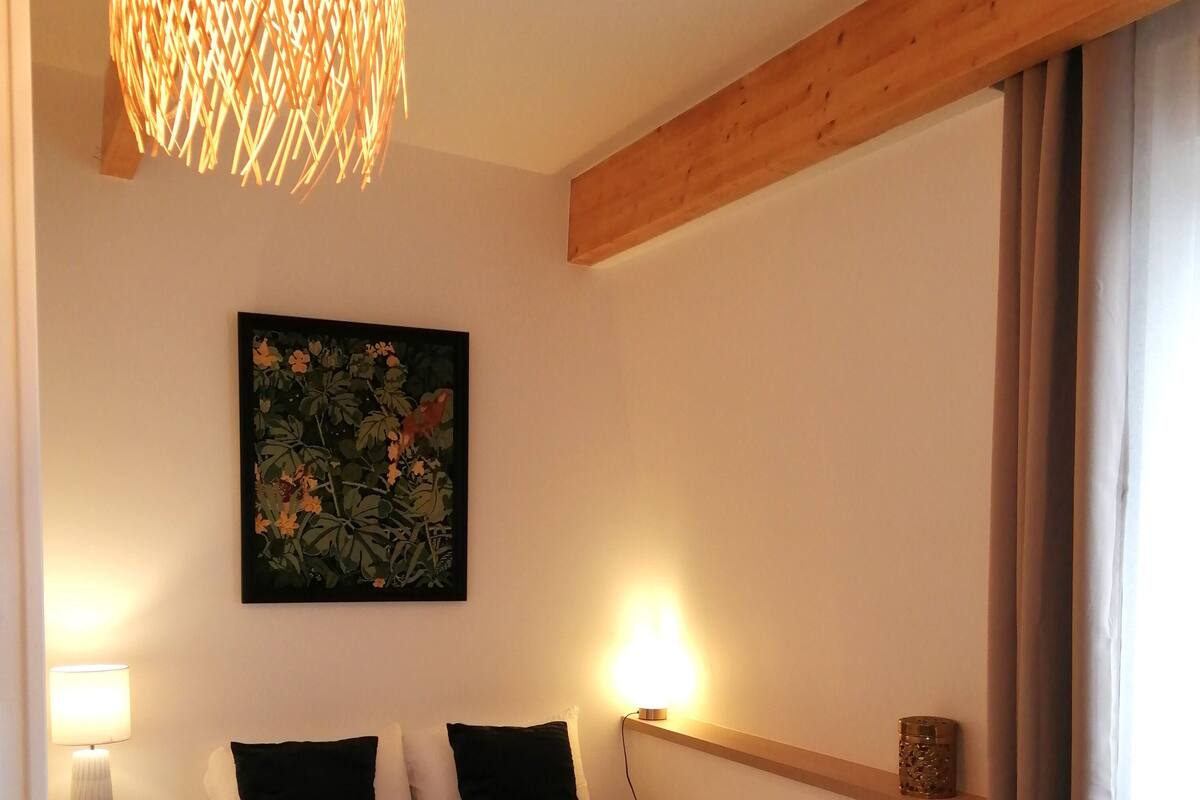A hotel room with a bed, a bedside table with a lamp, a small round table, a wall-mounted shelf, and a hanging light fixture.