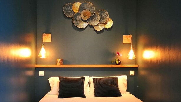 A hotel room with a bed, two black pillows, a wooden headboard, a wall-mounted decorative piece, and two wall sconces. - Studios confort  VILLA VOLTAIRE (Clamart)
