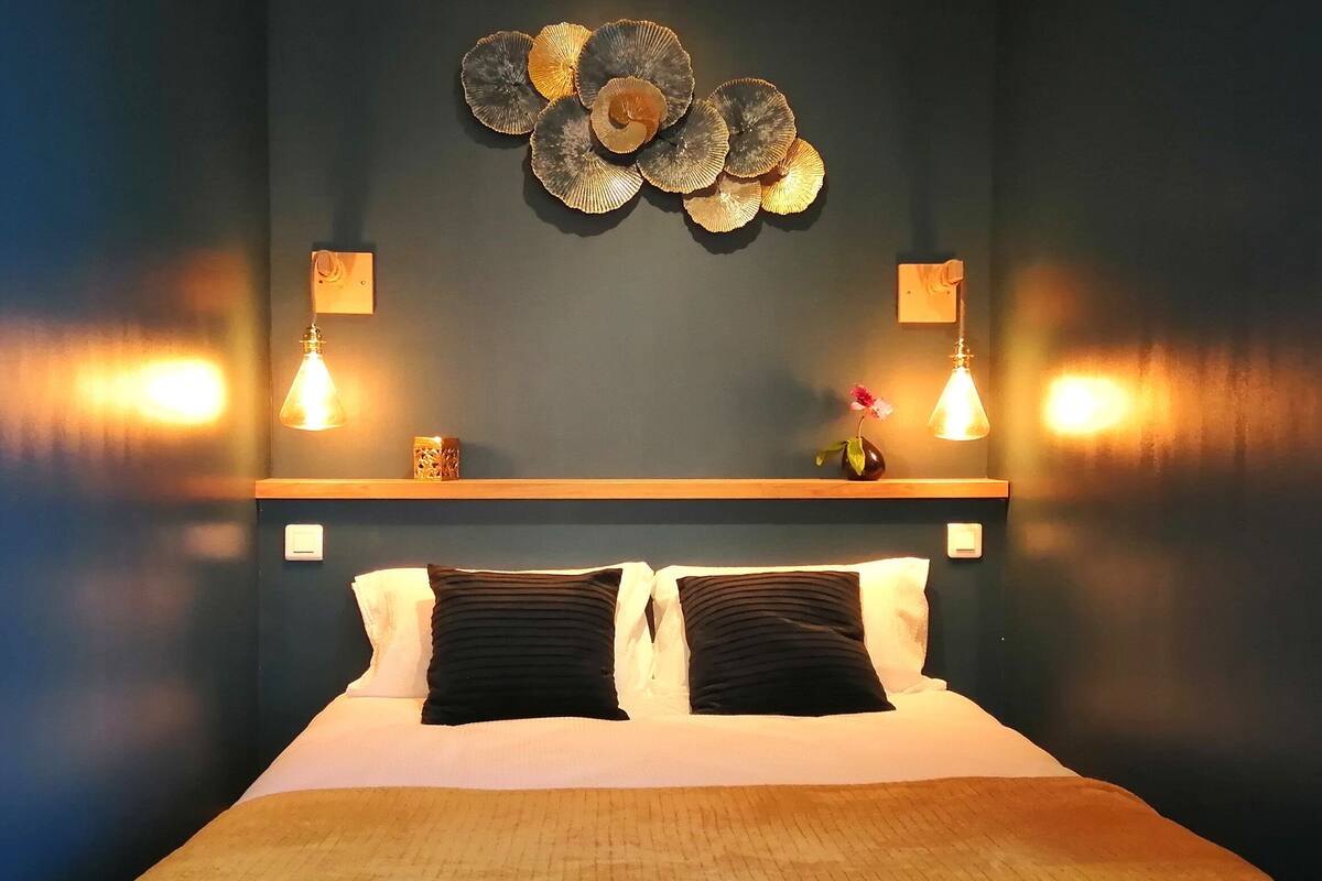 A hotel room with a bed, two black pillows, a wooden headboard, a wall-mounted decorative piece, and two wall sconces.