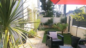 A patio area with outdoor furniture, including a chair and a table, surrounded by potted plants and a small lawn. - Studios confort  VILLA VOLTAIRE (Clamart)