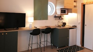 A compact kitchen with a TV, two bar stools, a microwave, a sink, and a hanging light fixture. - Studios confort  VILLA VOLTAIRE (Clamart)