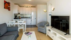 Ein modernes Wohnzimmer mit grauem Sofa, einem weißen Couchtisch, einem grauen Sessel, einem Flachbildfernseher auf einem weißen Schrank und einer Küche im Hintergrund.