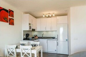 Une cuisine compacte avec des meubles blancs, un four à micro-ondes, un réfrigérateur et un coin repas avec des chaises blanches.