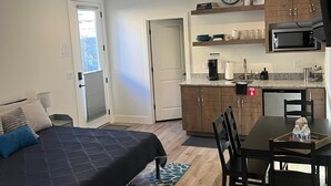 A bedroom with a bed, a desk, and a chair. There is a kitchenette with a microwave, sink, and cabinets. A clock is on the wall. - Cozy Casita by Sand Hollow and Zion (Hurricane)