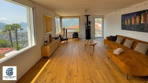 A spacious living room with wooden flooring, a sofa, a coffee table, and a view of a cityscape through large windows. - Casa Teatro Panoramico (Locarno)
