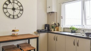 Eine Küche mit Holztisch, zwei Hocker, einer Uhr, einem Fenster und einem Waschbecken.