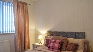 Ein Schlafzimmer mit einem Bett, einem Nachttisch, einer Lampe und einem Fenster mit Vorhängen.