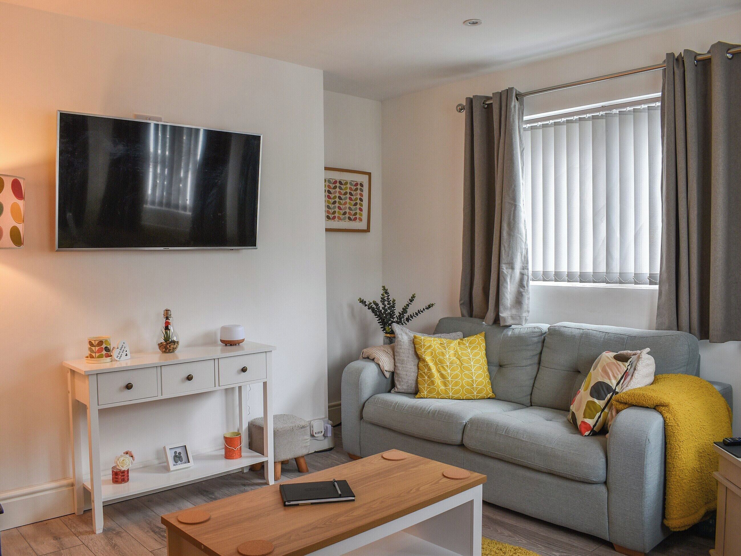 A modern living room with a grey sofa, a wooden coffee table, a flat-screen TV, a white console table, and a window with curtains.