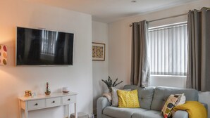 Ein modernes Wohnzimmer mit einem grauen Sofa, einem Holztisch, einem Flachbildfernseher, einem weißen Konsolentisch und einem Fenster mit Vorhängen.