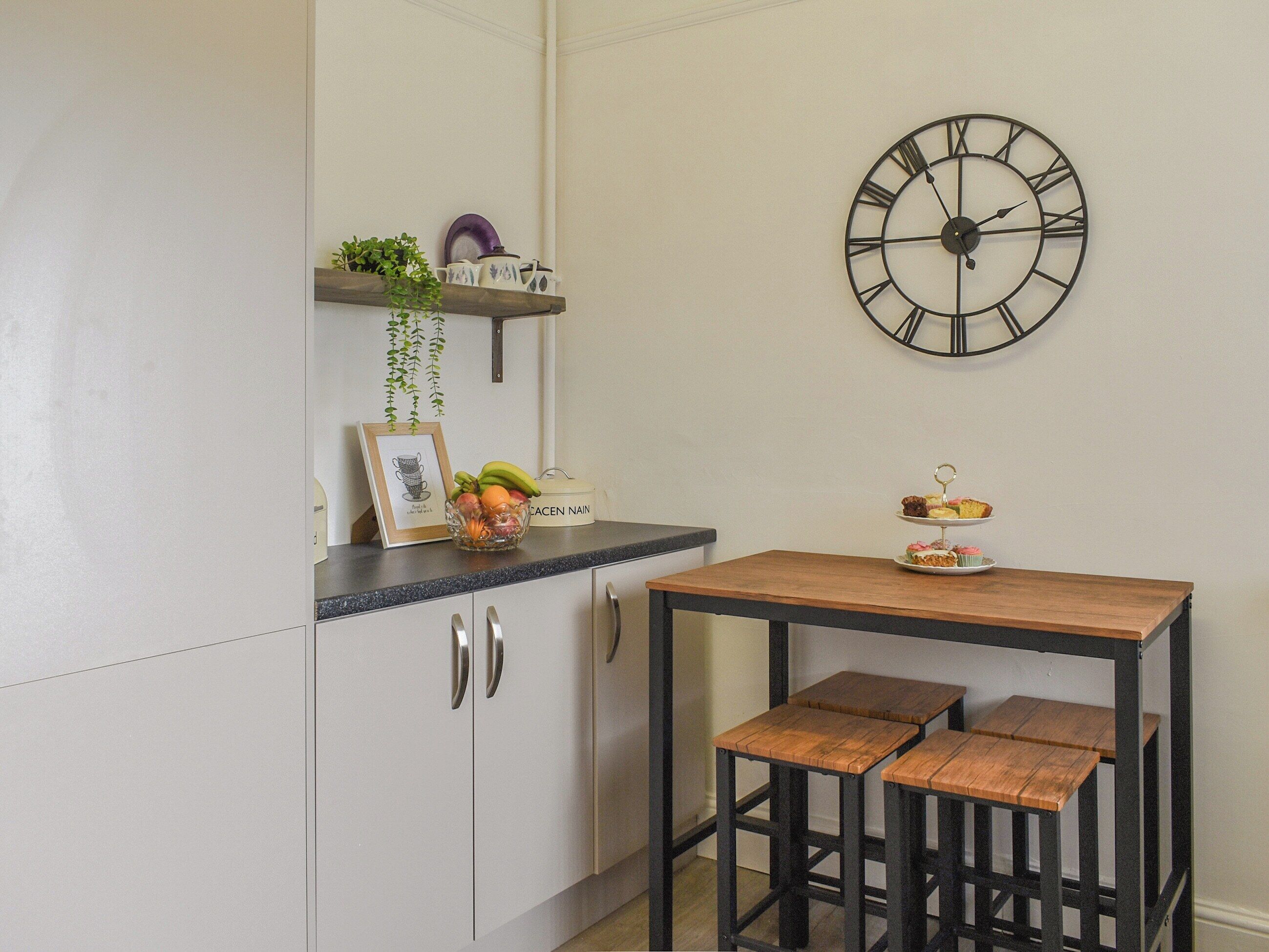A kitchen with white cabinets, a black countertop, a wooden table with stools, a wall clock, and a shelf with kitchenware and a plant.