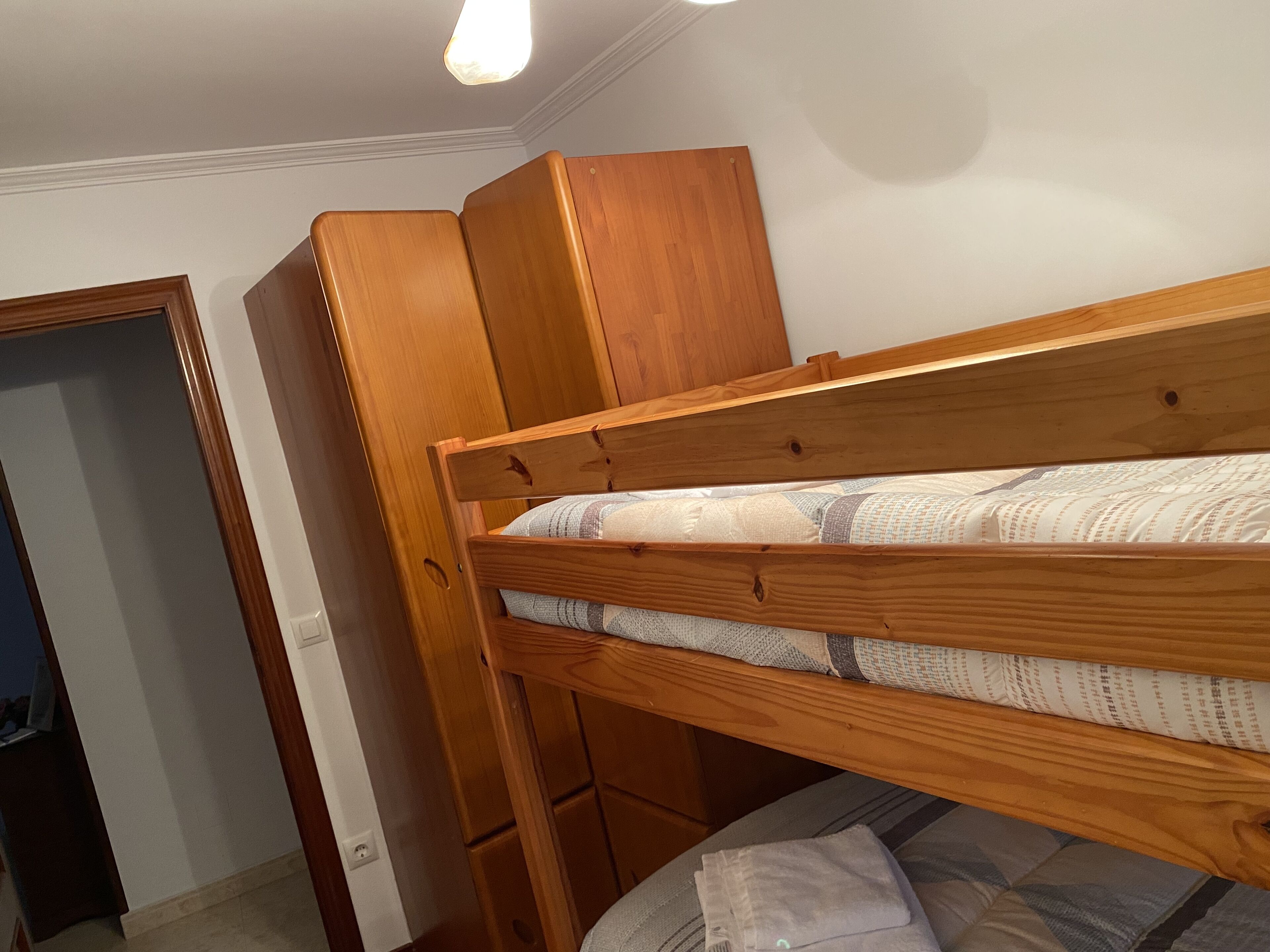 A bunk bed with a wooden frame, a mattress with patterned bedding, and a wooden wardrobe.