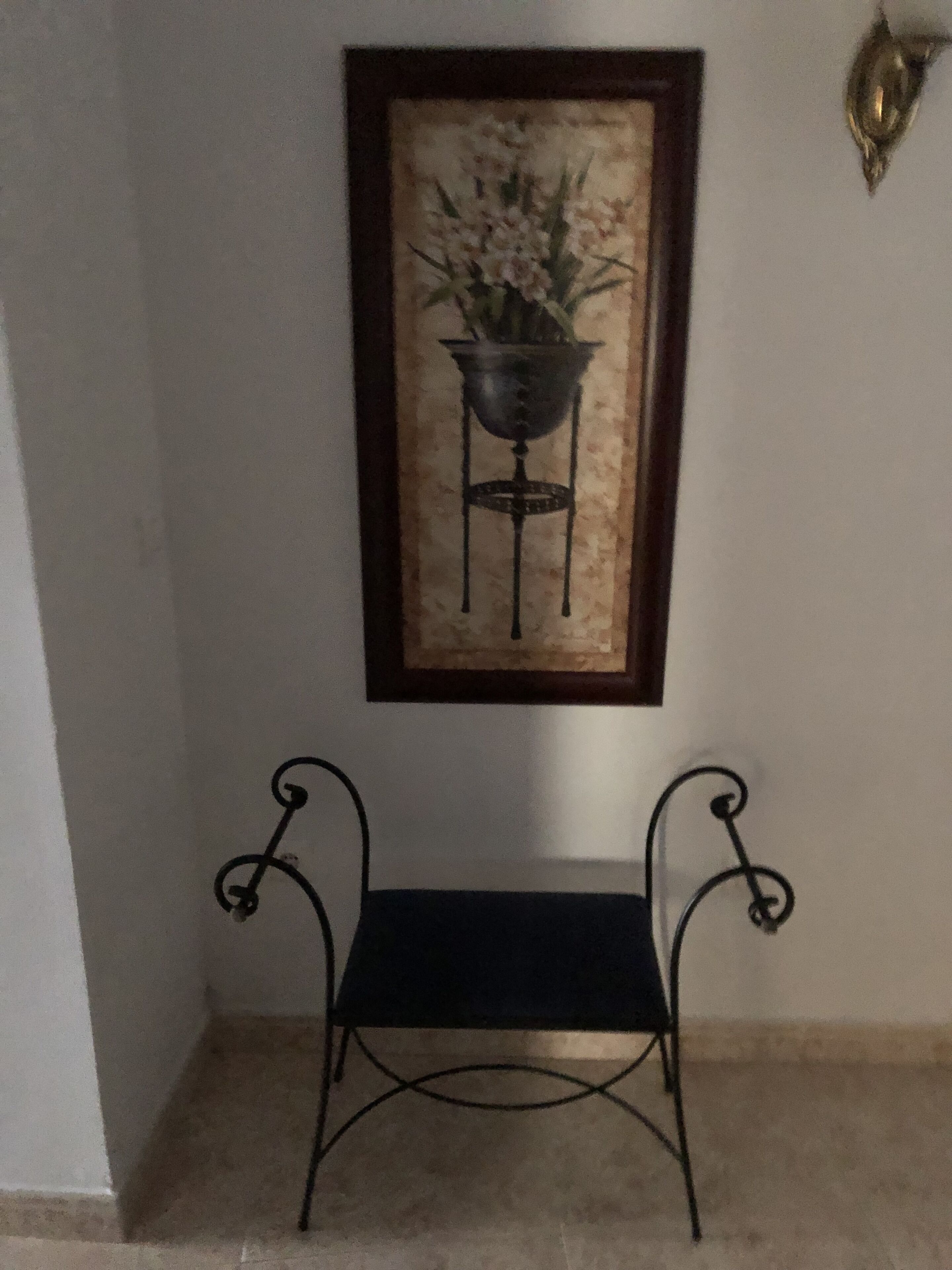 Un cuadro enmarcado que muestra una maceta sobre un pedestal, un banco pequeño de color negro y una lámpara fijada a la pared.