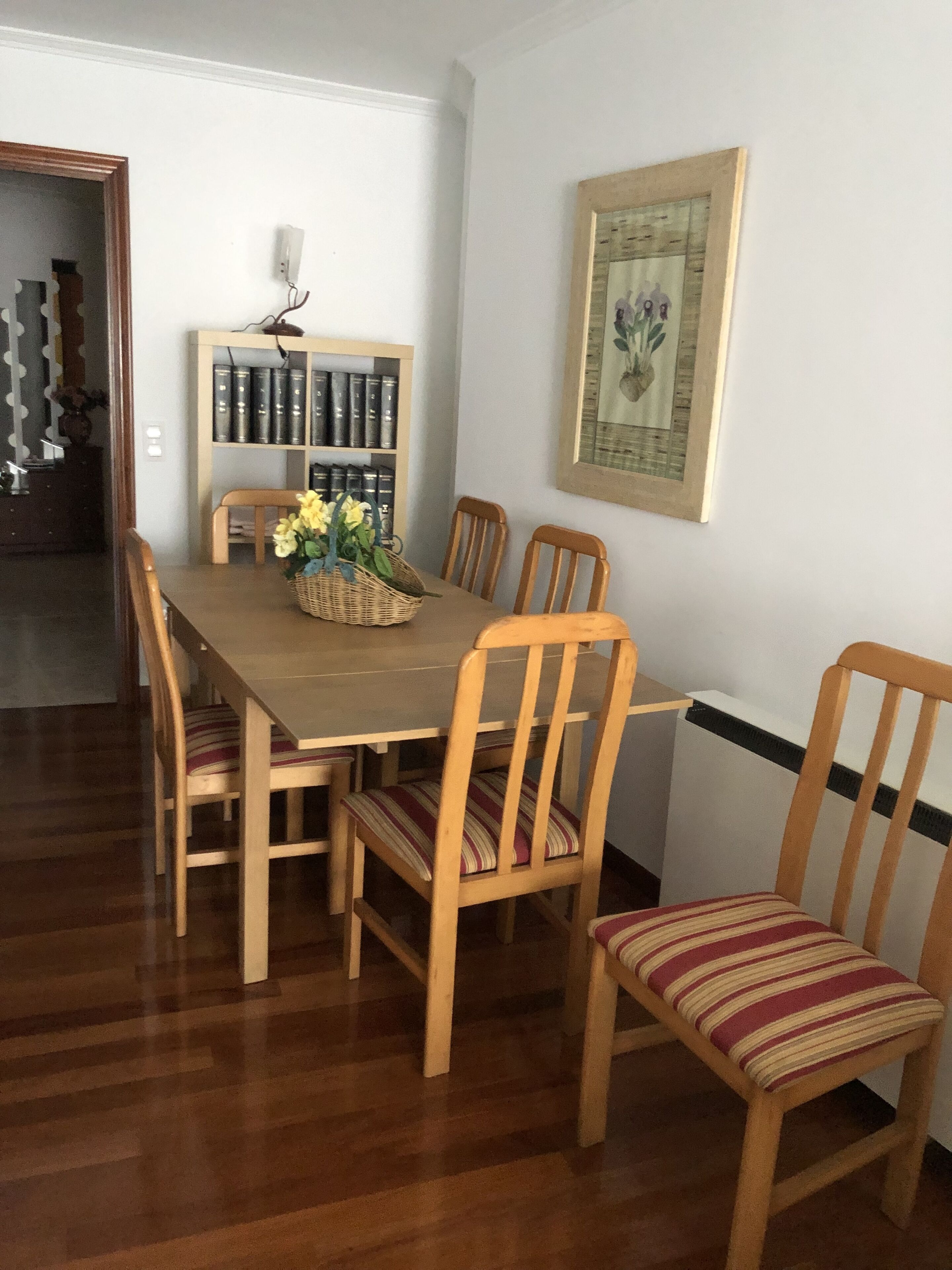 A dining area with a wooden table and chairs, a bookshelf, and a framed picture on the wall.