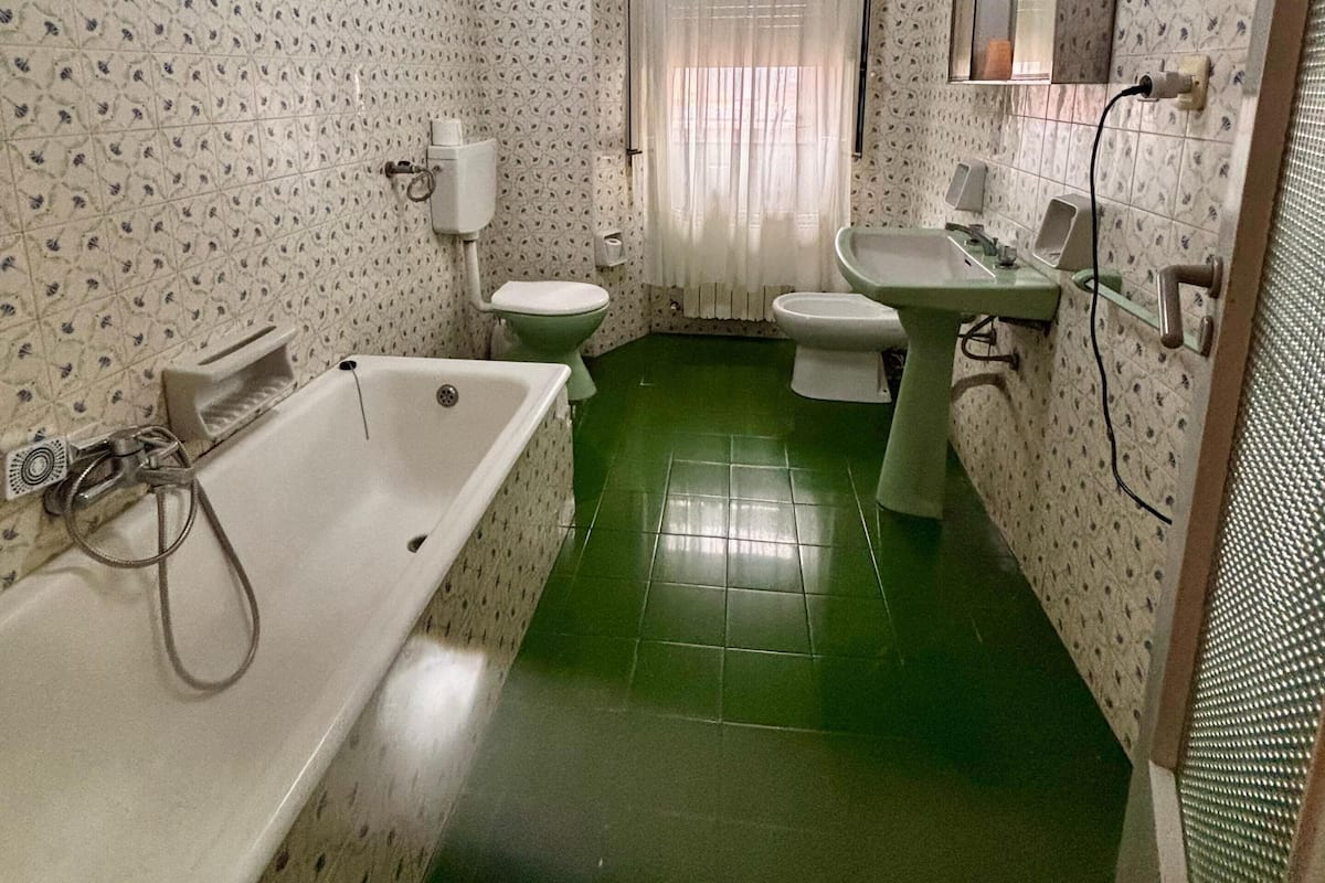 A bathroom with a green floor, white patterned walls, a white bathtub, two toilets, a sink, and a mirror.