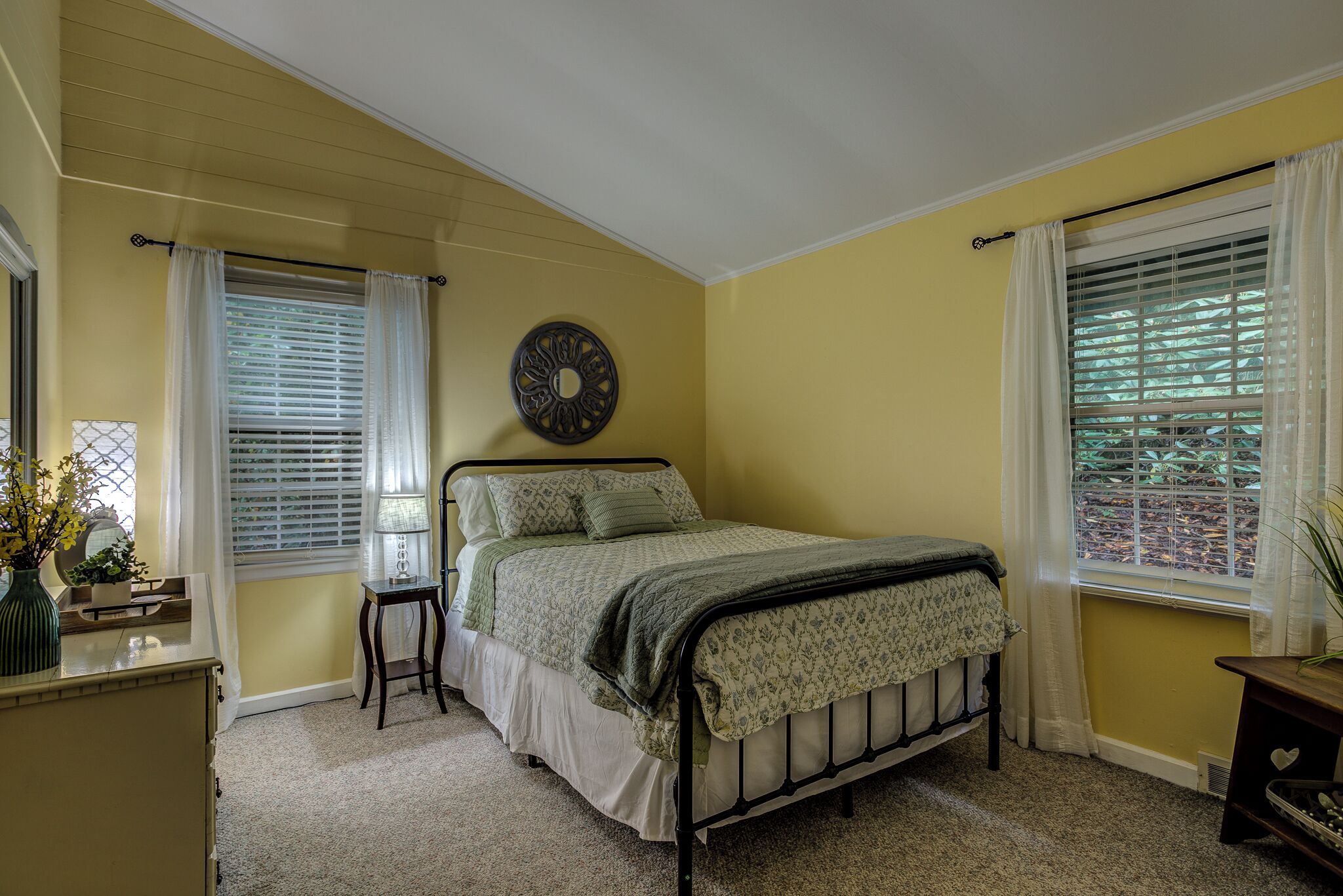 A bedroom with a bed, a bedside table, a lamp, a decorative wall piece, and a window with blinds.