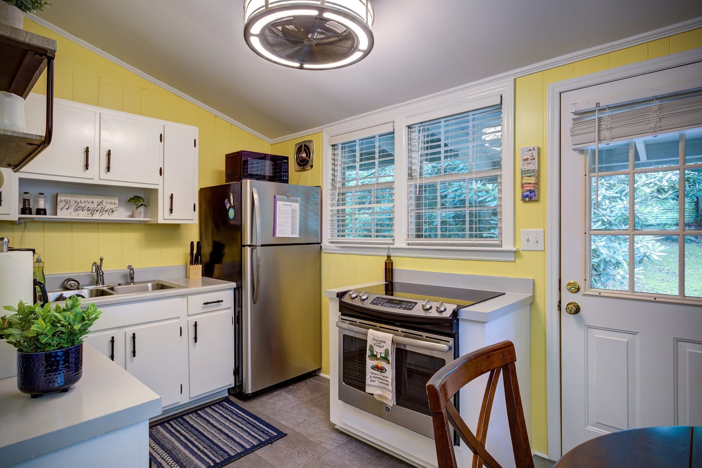 A kitchen with white cabinets, a stainless steel refrigerator, a built-in oven, and a window with blinds.
